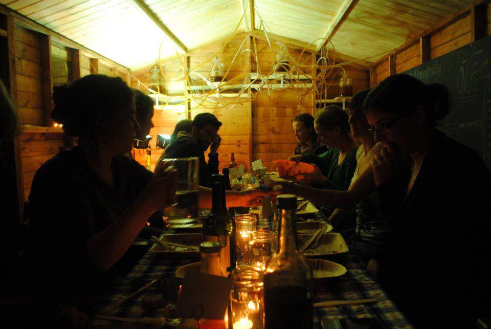 People gathered in a shed with pipes and jars of liquid in the ceiling, and food spread across the table in front of them.