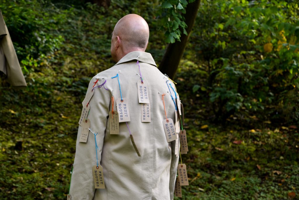 A man wearing a coat with labels reading "Fruit Routes" attached with safety pins and string