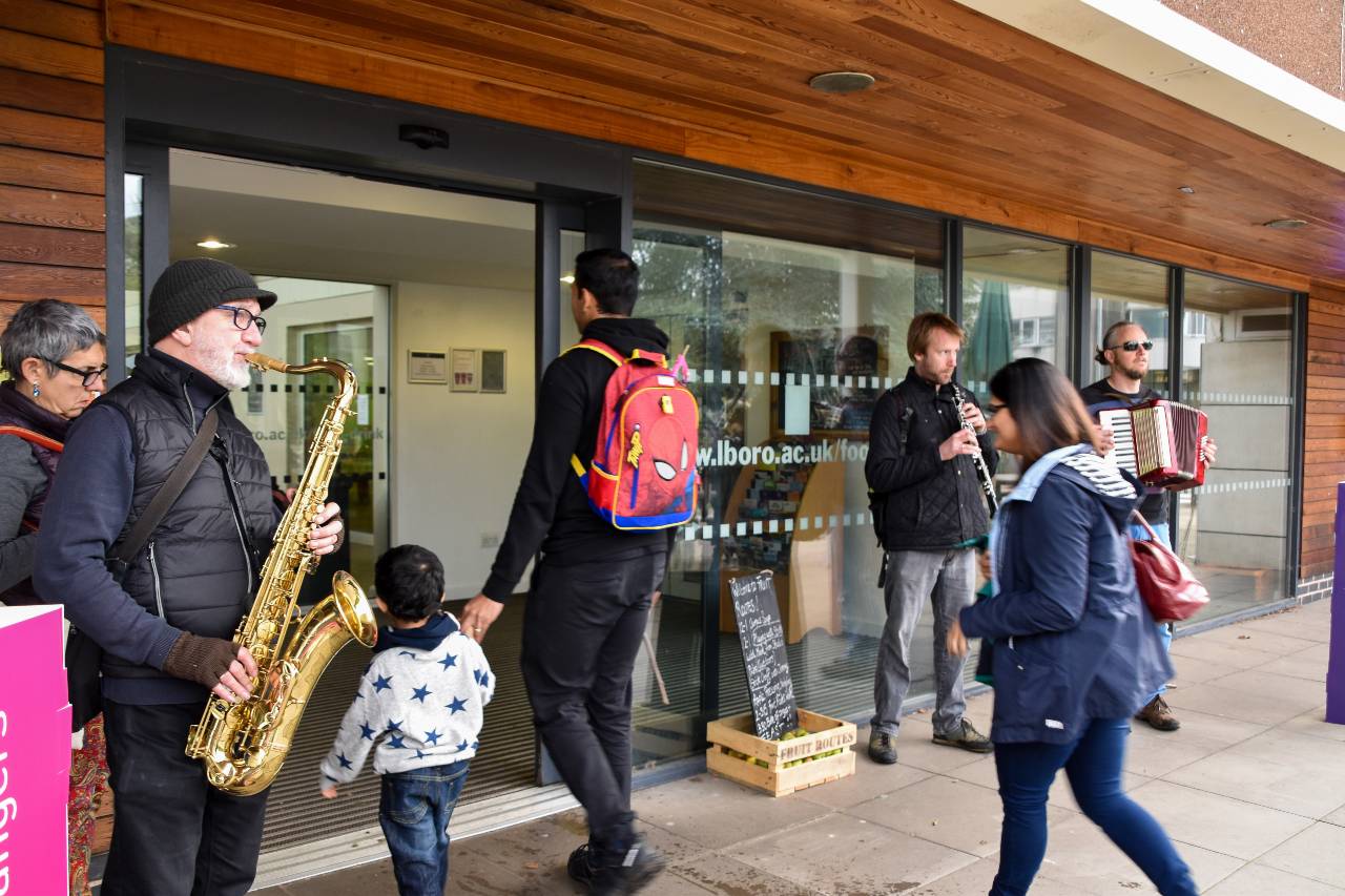 3 musicians are stood in front of glass windows where a family walks through an automatic door.