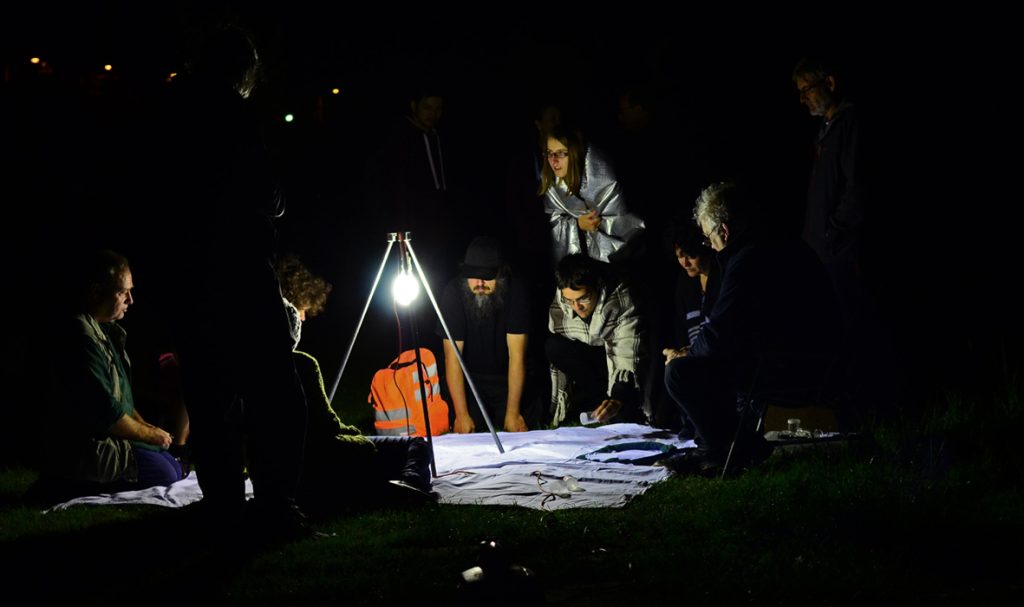 A group of people gathered in the dark around a lamp shone on a blanket on the floor.