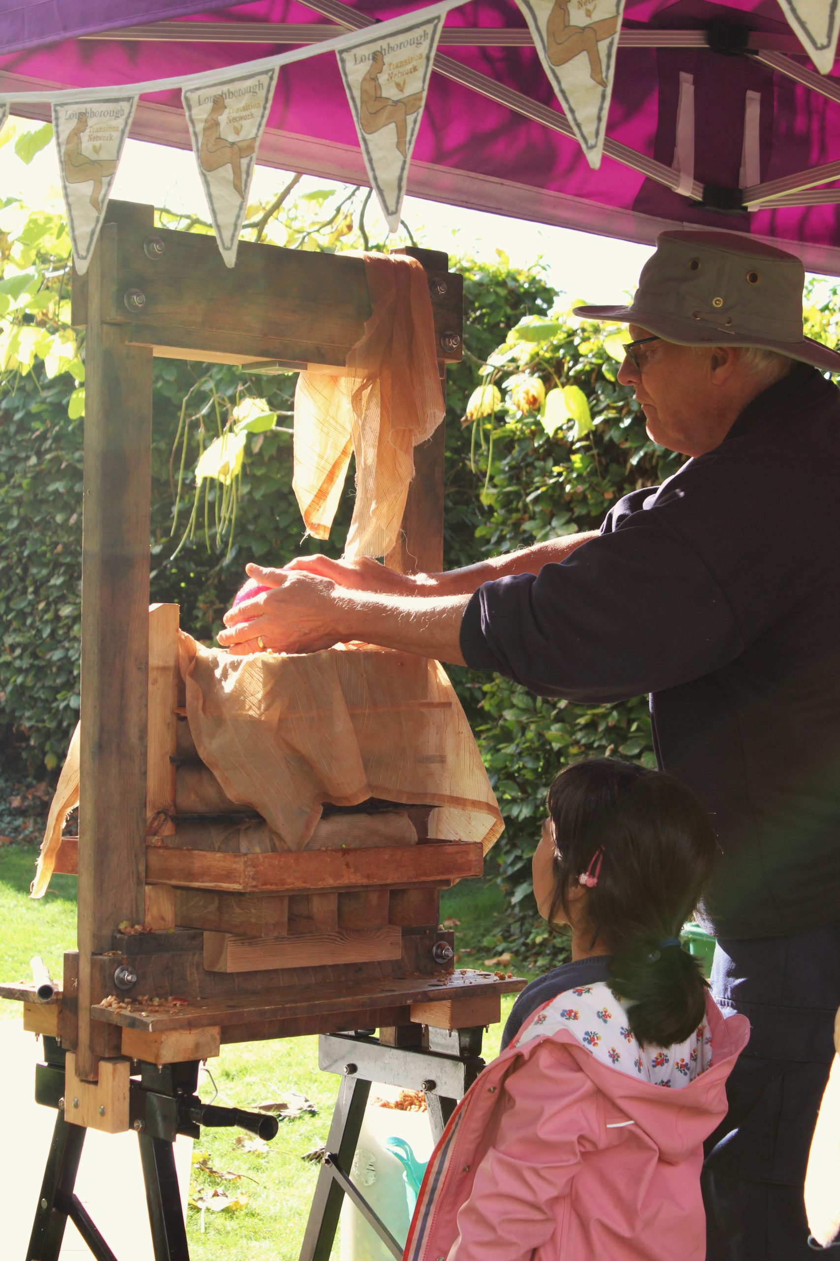 A male adult feeding apples into a wooden apple press outside while a young child looks on